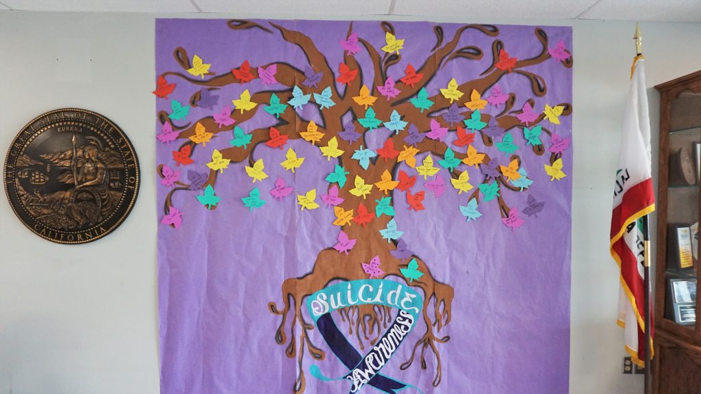 A painting of a tree with messages on the leaves and a banner that reads: Suicide Awareness.