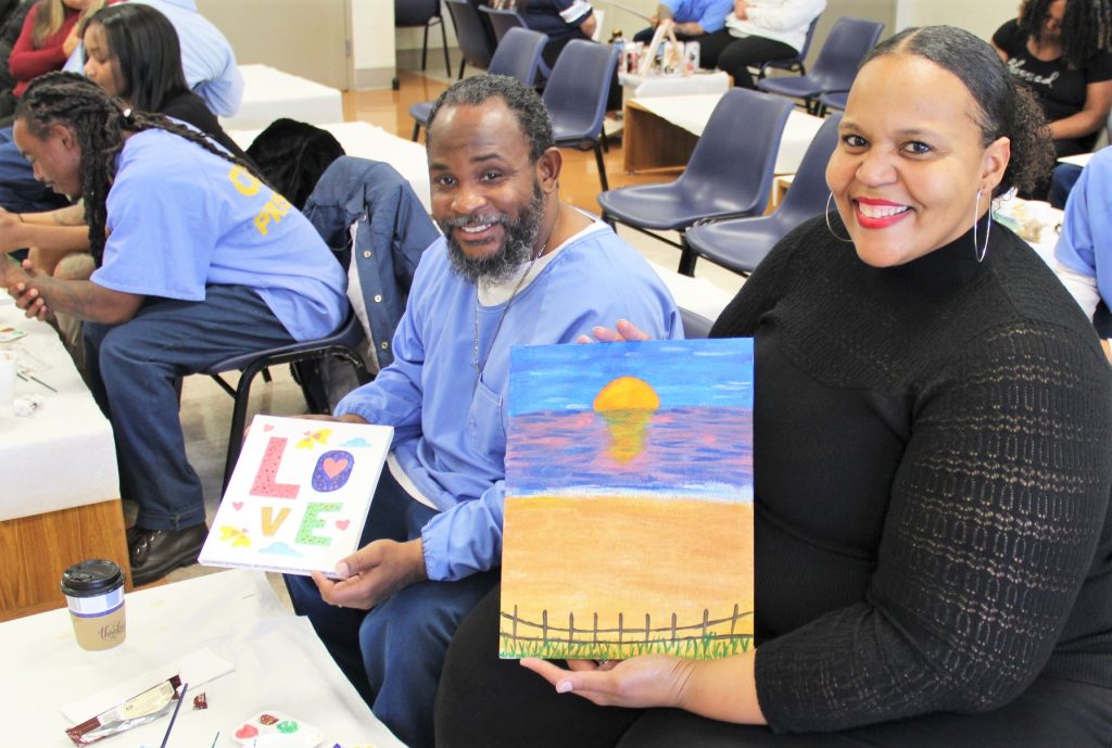 Smiling people hold up their paintings at a visiting event inside a correctional facility.