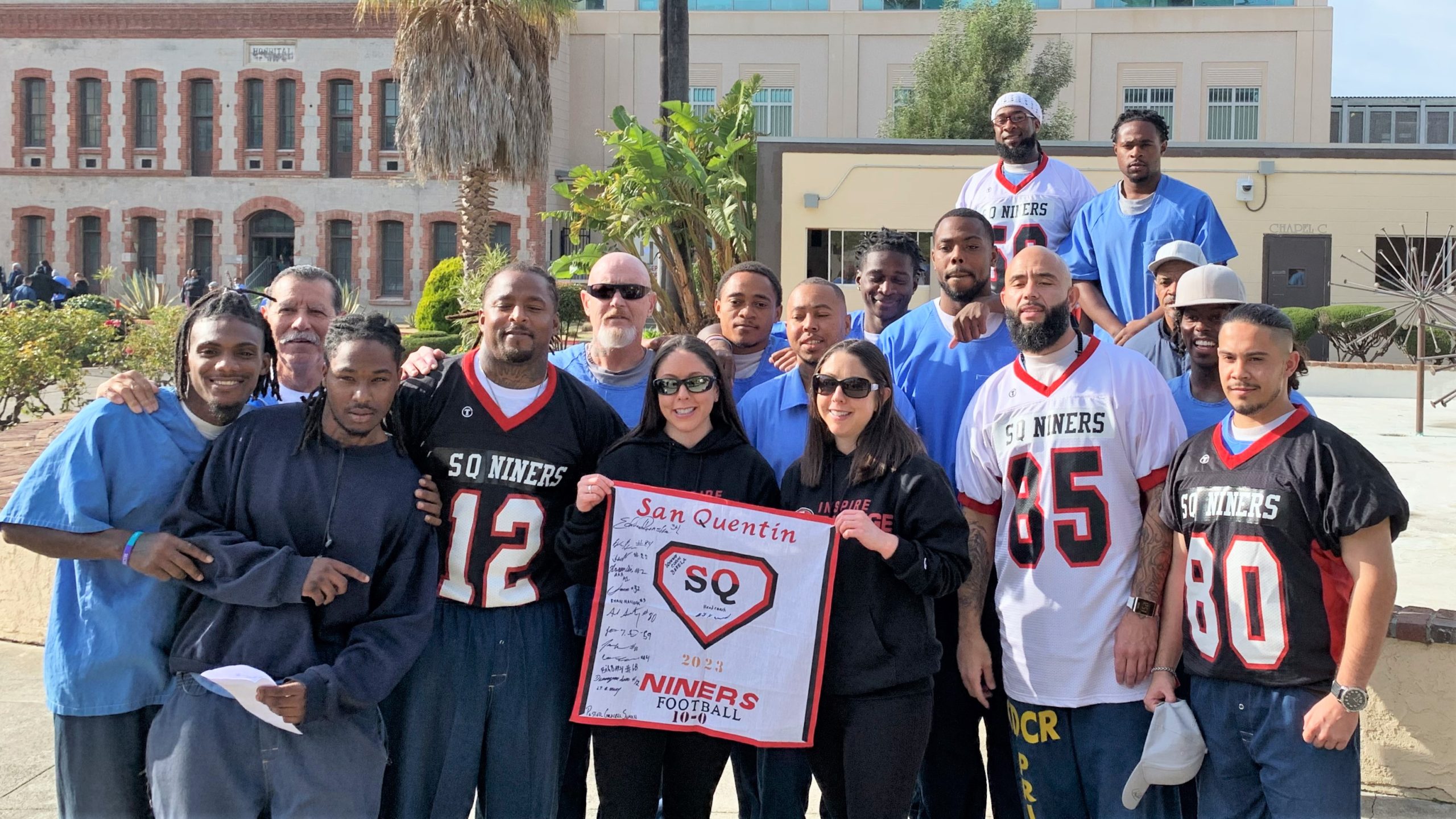 A group of people, some wearing football jerseys and holding a San Francisco 49ers sign, pose for a photo with incarcerated people inside San Quentin Rehabilitation Center.