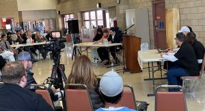 Valley State Prison students and university students at tables prepare for a debate in front of cameras and an audience.