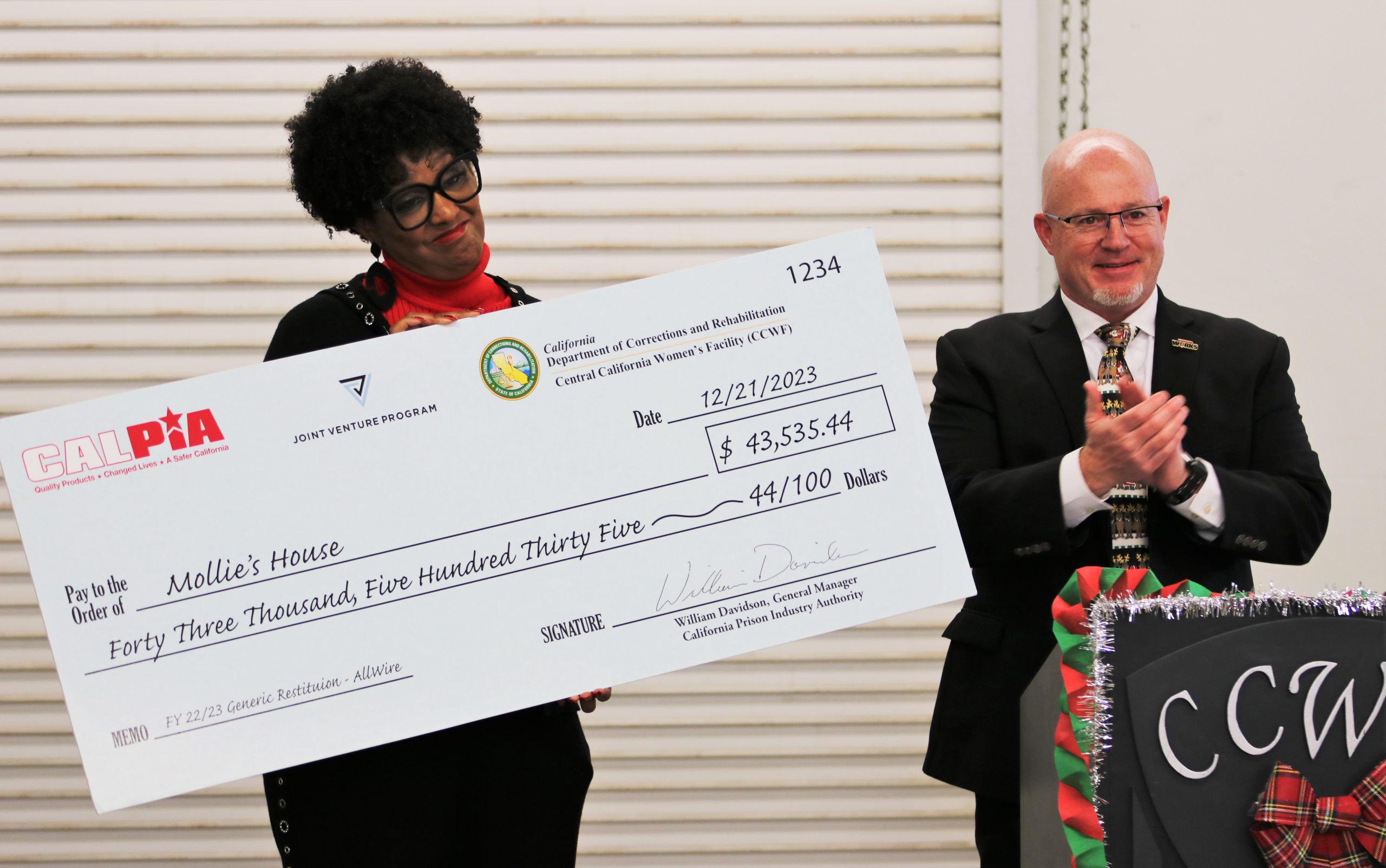 A woman holds an over-sized donation check presented by CALPIA as another person applauds.