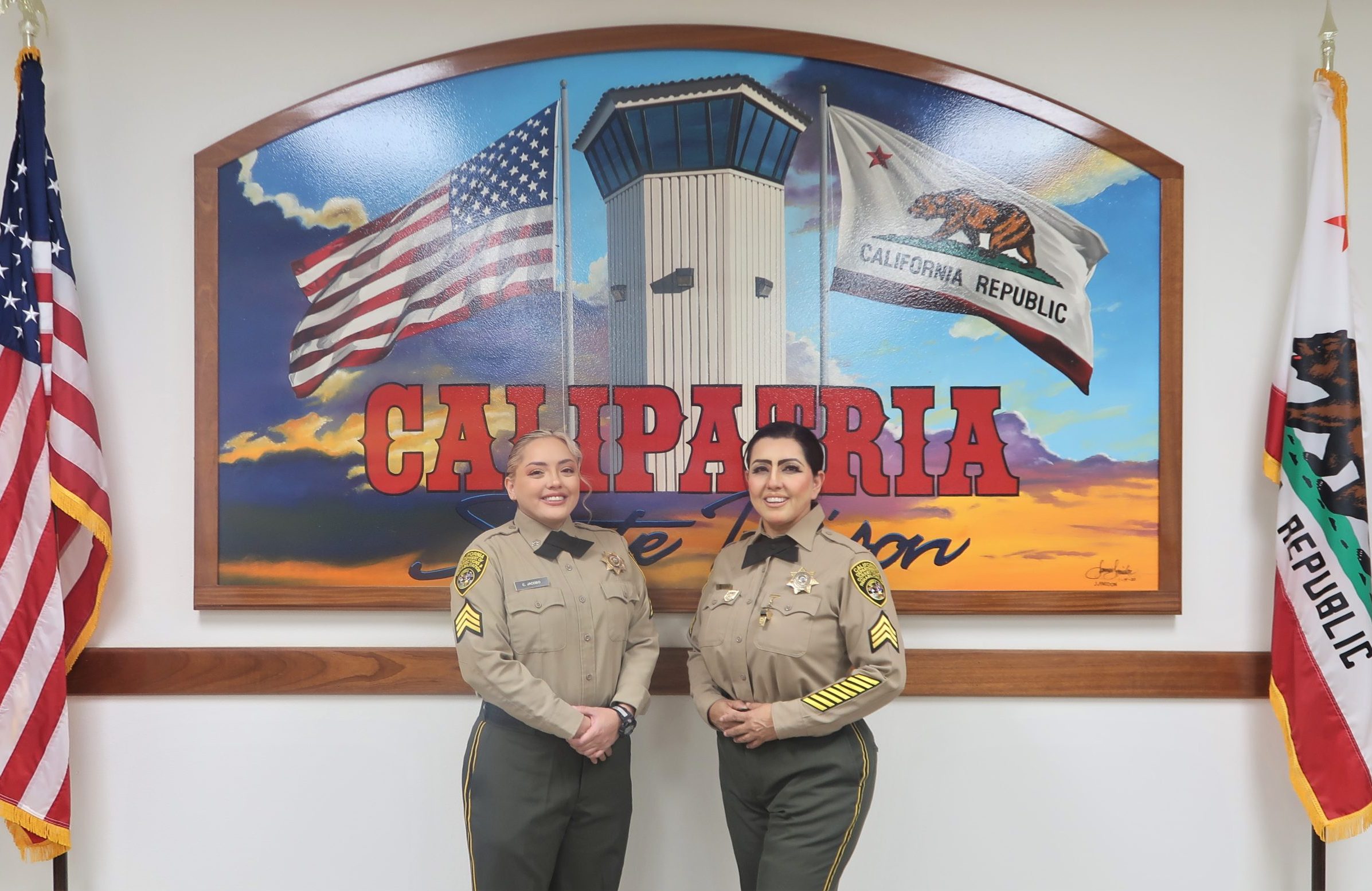 Female correctional sergeants, a daughter and her mother, stand in front of a mural of Calipatria State Prison.