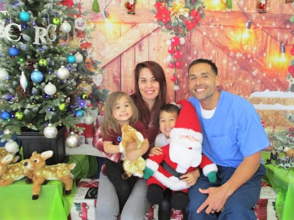 Two young children, their mother and an incarcerated person pose next to a Christmas tree and a holiday themed backdrop.