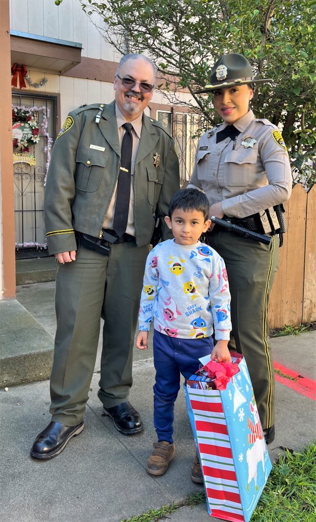 Two CDCR staff wearing uniforms stand beside a child holding a gift bag.