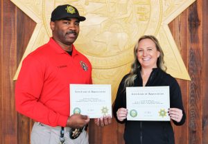 Two recruiters of the month hold certificates of appreciation while standing in front of the California Department of Corrections and Rehabilitation seal.