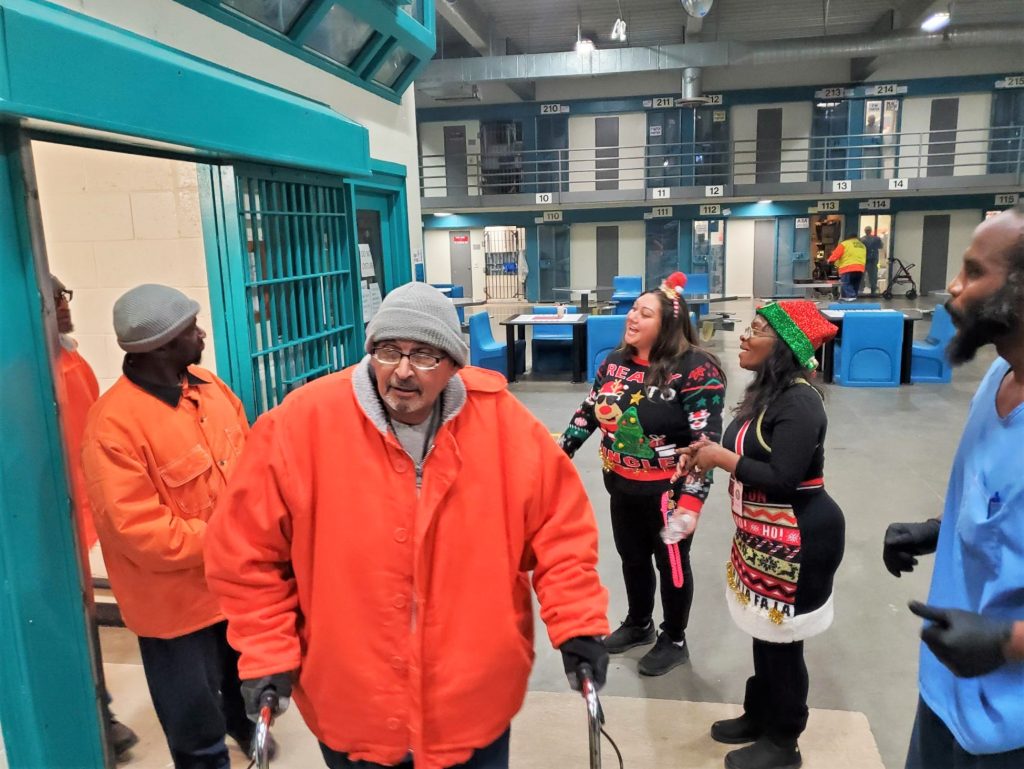 Incarcerated people enter a prison housing unit, greeted by people in holiday attire.