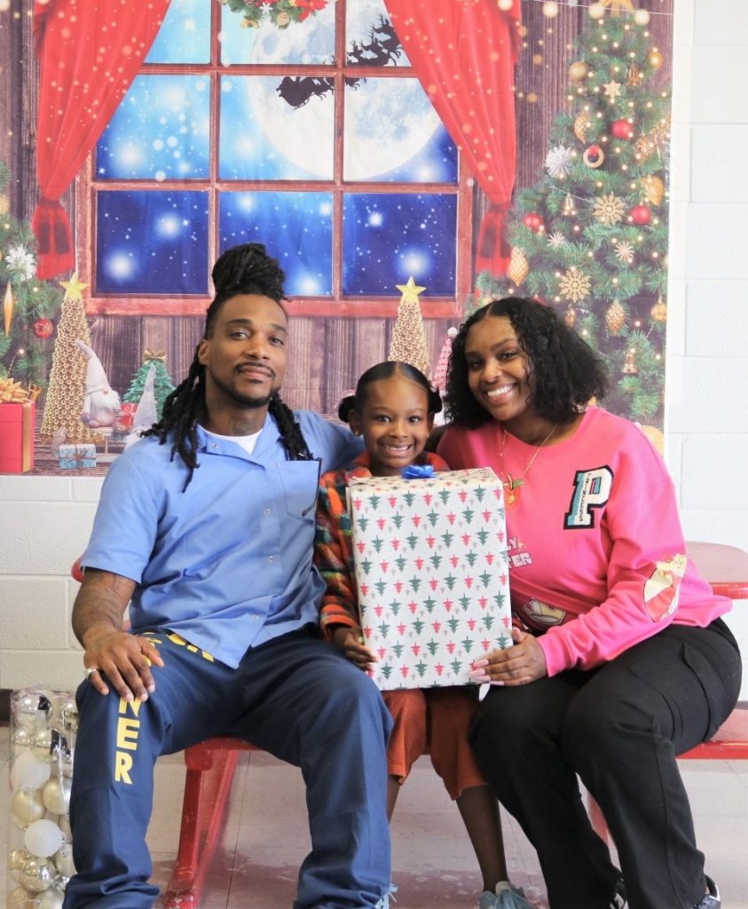 An incarcerated man sits in front of a Christmas backdrop with his child and child's mother.