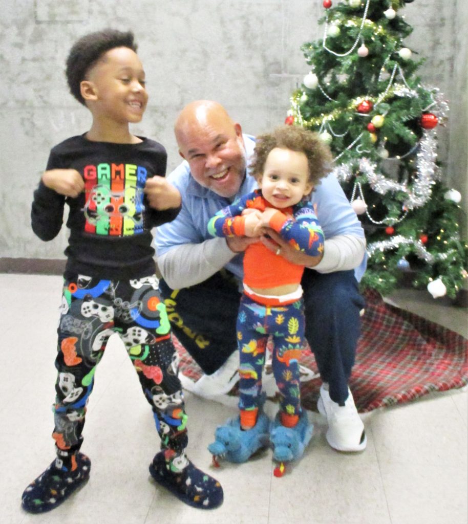Two children wearing pajamas play with their incarcerated loved one at California State Prison Sacramento. A Christmas tree can be seen in the background.