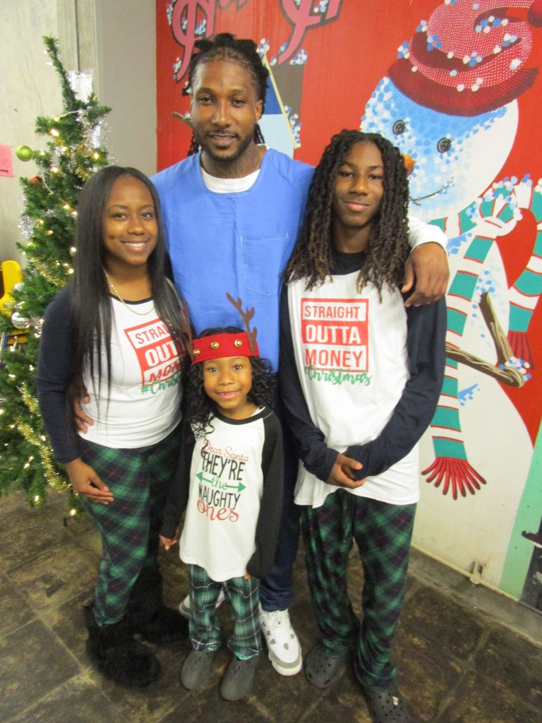 Three people wearing holiday pajamas and an incarcerated person standing beside a Christmas tree.