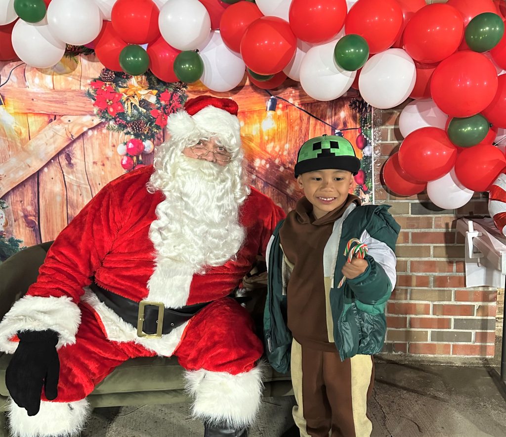 Santa sitting in a chair with a photo backdrop behind him and a child smiles while holding candy canes.
