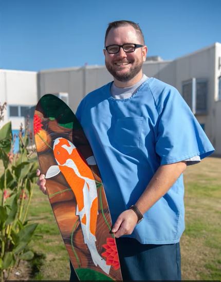 A man in blue holds a skateboard painted with a koi fish