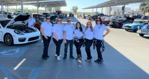 Six people in white tee-shirts stand in front of a parking lot full of classic cars.