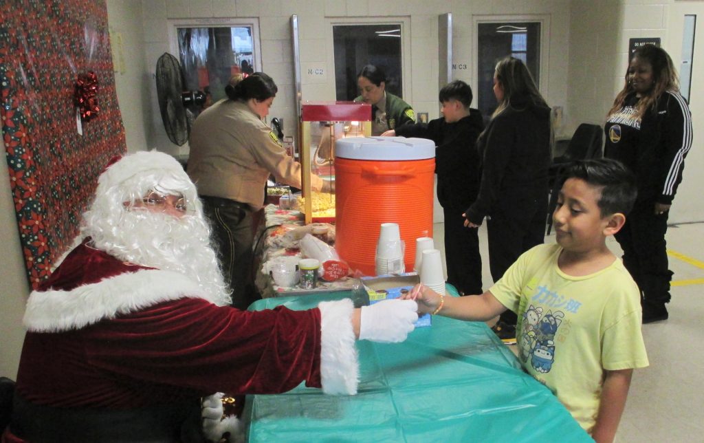 Santa hands a candy cane to a child in a prison visiting room.