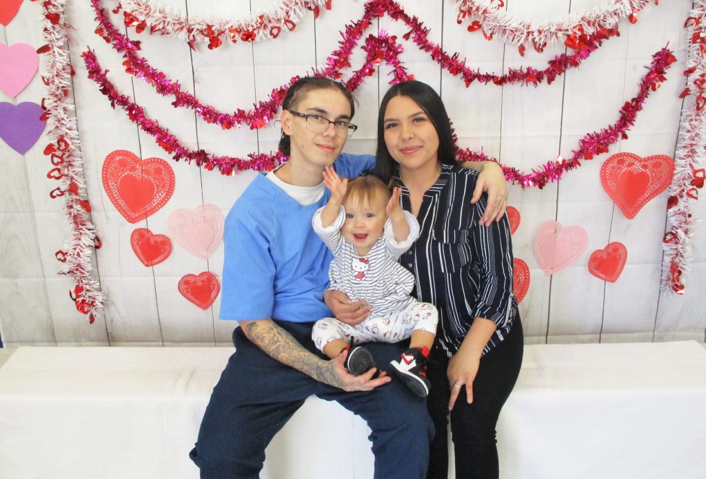 A couple, one of them incarcerated hold a baby for a Valentine's photo against a decorated backdrop.