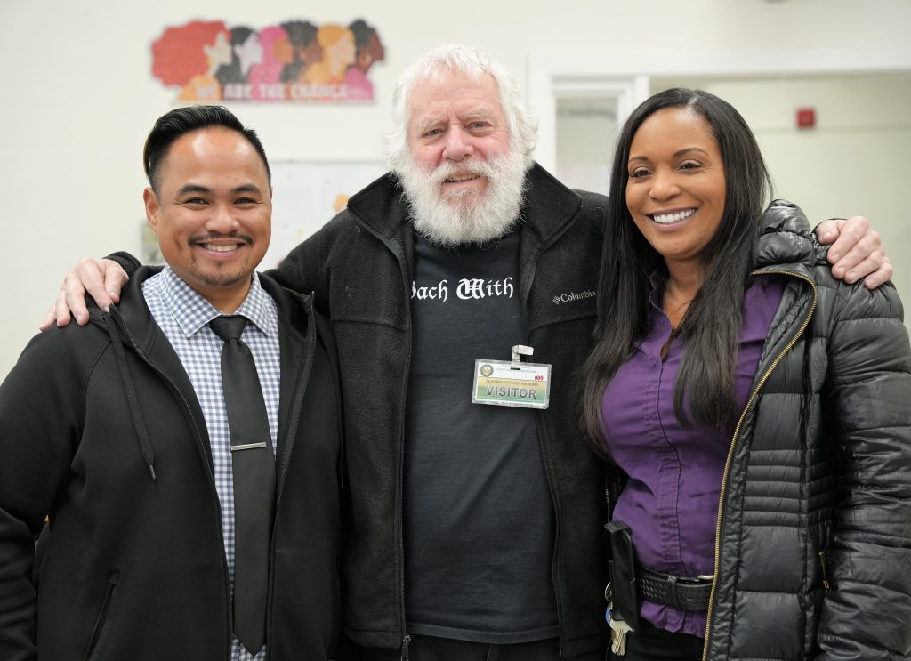 Two prisons staff people standing in a library with a man wearing a Bach with Verse t-shirt.