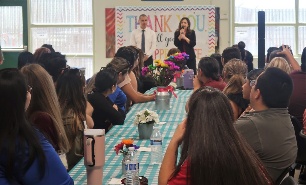 Two prison leaders speak to staff who are seated at a checkered tablecloth covered table.