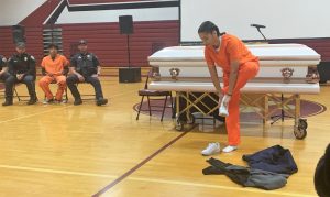 An incarcerated female firefighter stands in front of a casket in a high school gymnasium.