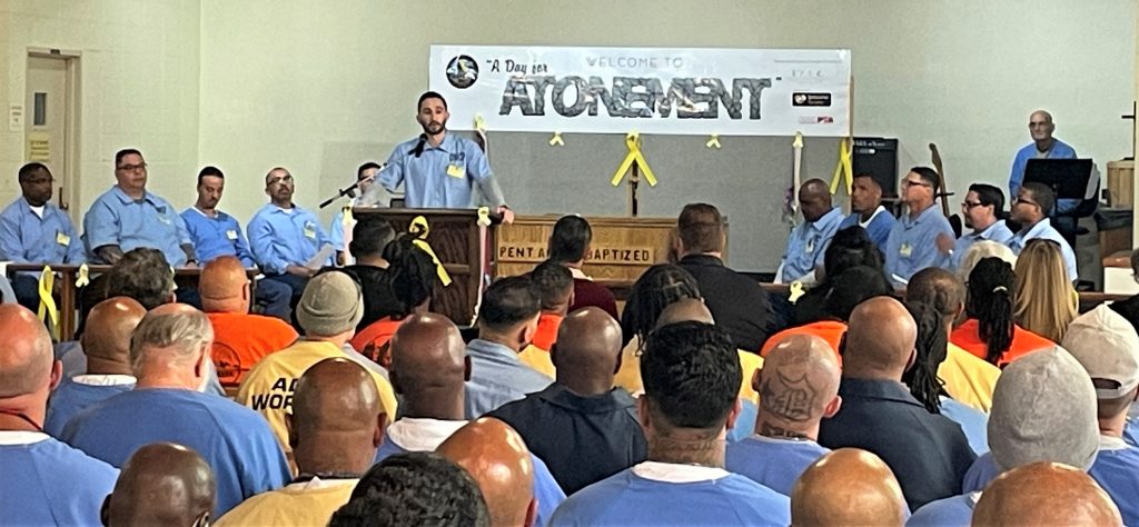 An incarcerate person speaks in front of a large gathering with a banner behind him that reads "A Day for Atonement."