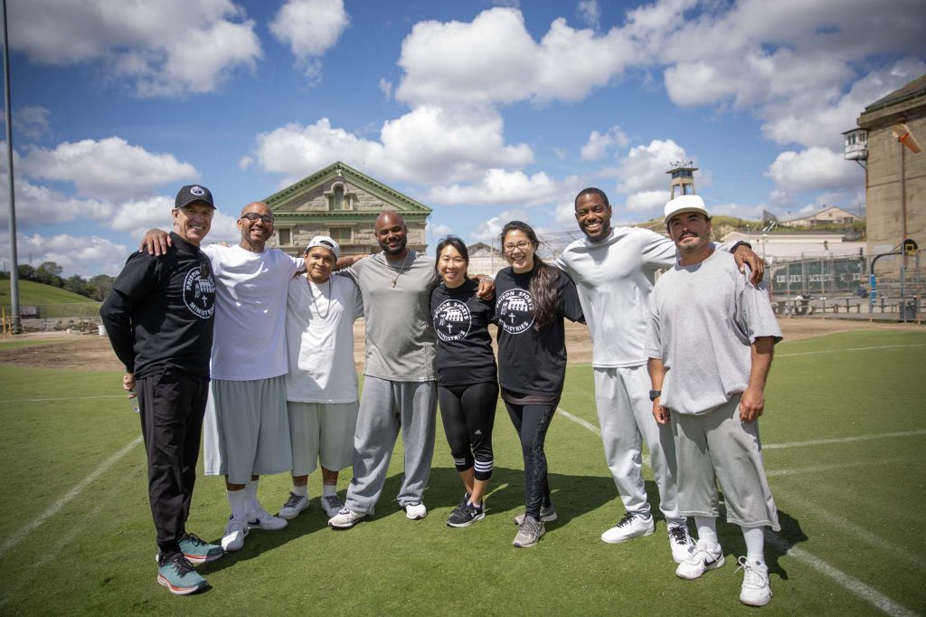 Cloudy sky with a chapel in the background at Folsom prison. A group of volunteers and incarcerated people stand on a field.