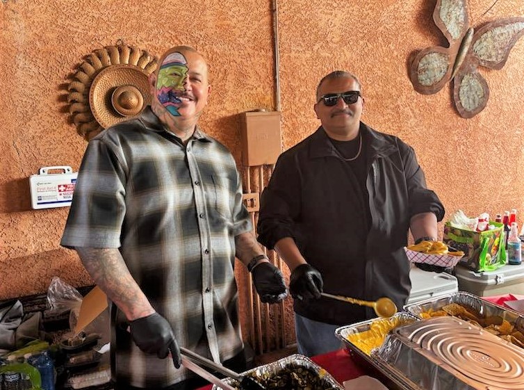 Two men serve food during an event. One man has an Easter egg painted on his face.