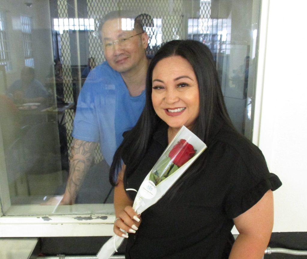 A man behind security glass and a woman holding a rose.