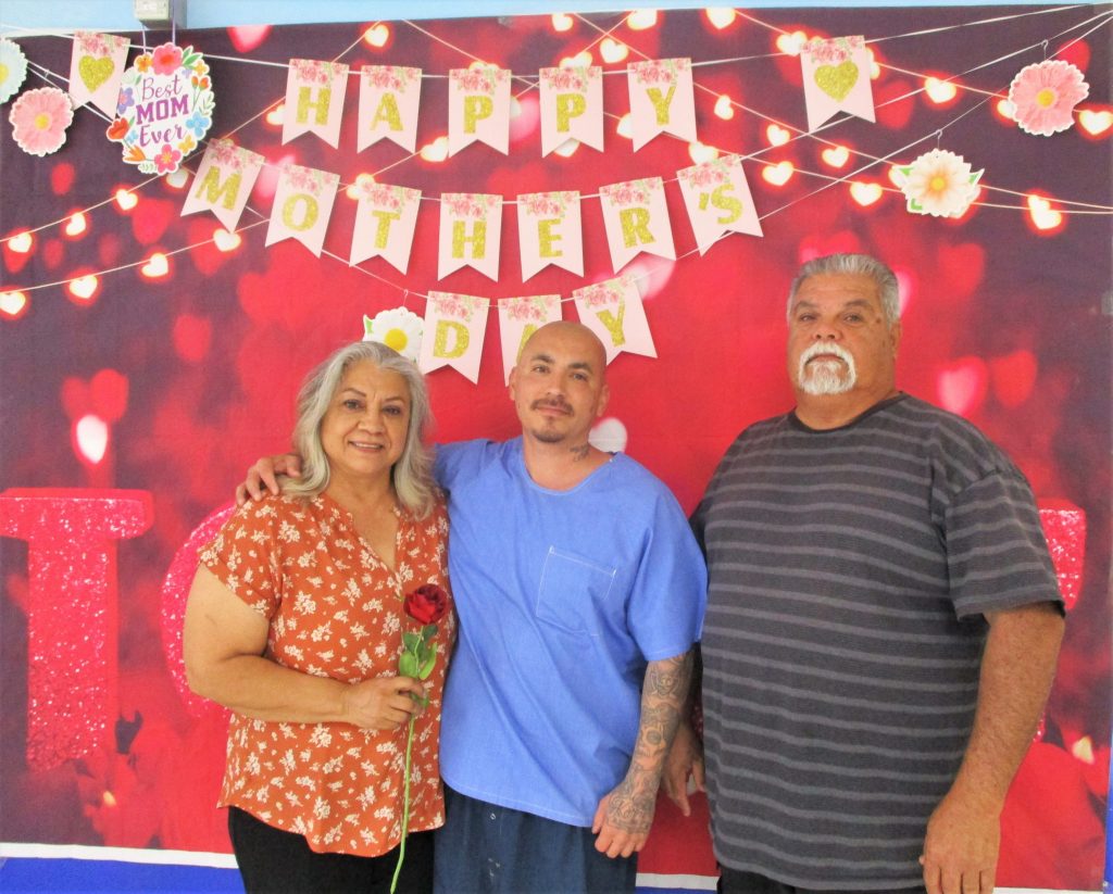 A mom and dad visit their incarcerated son. Behind them is a happy Mother's Day banner. 
