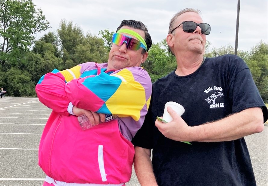 Two men, one wearing a colorful 1980s style track suit, strike a silly pose.