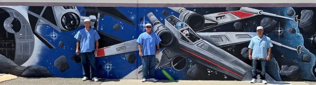 Star Wars paintings on a prison wall with three incarcerated artists standing in front.