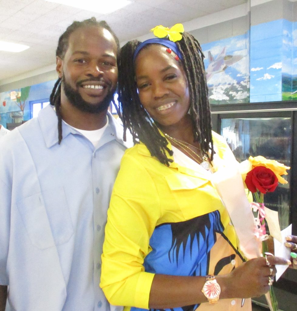 A woman holds a rose and standing beside an incarcerated man.