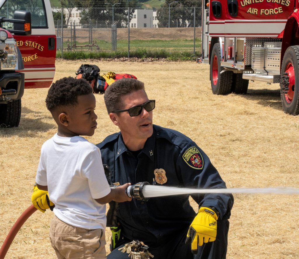A child holds a fire hose as an Air Force firefighter looks on. 