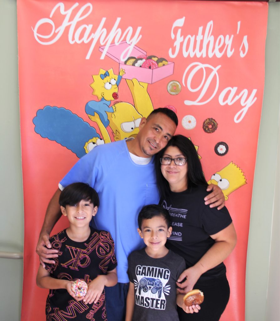 Donuts with Dads at California Health Care Facility, or CHCF, visiting with two kids holding donuts, their incarcerated father, and their mom in front of a Simpson's Happy Father's Day banner.