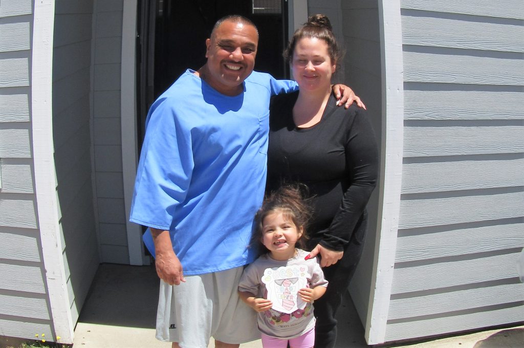An incarcerated man has his arm around a woman as they stand with a small child to pose for a family photo.