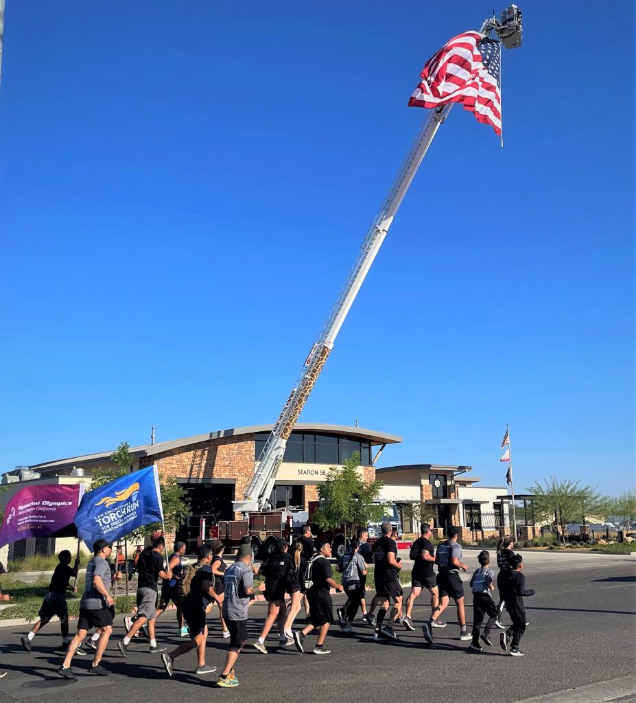 Law Enforcement Torch Run passes a fire department with American flag flying overhead.