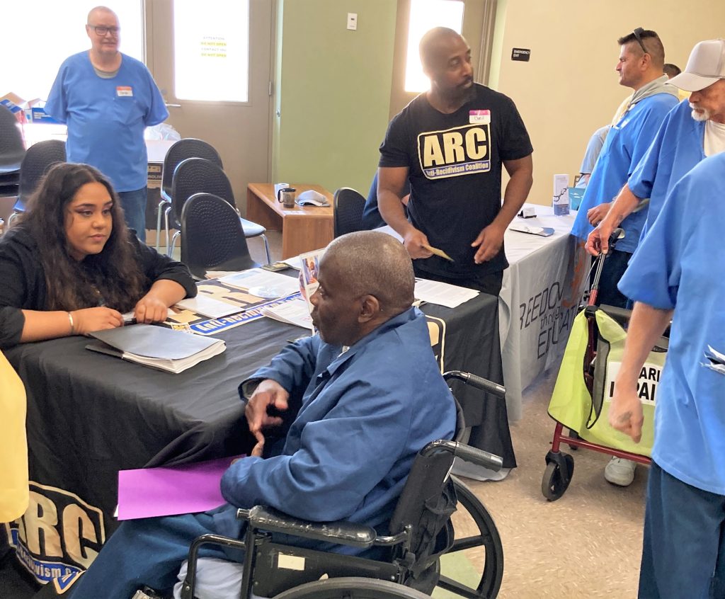 A man wearing a t-shirt with the letters ARC, which stands for Anti-Recidivism Coalition, speaks to incarcerated people at a CHCF reentry resources fair. 