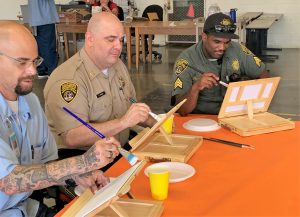 A correctional captain, sergeant and an incarcerated person sit together at a table to paint at California Medical Facility (CMF).