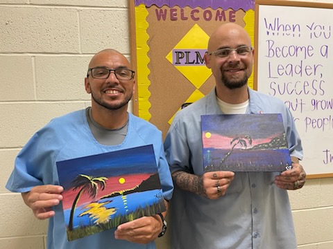 Two incarcerated people holding paintings. 