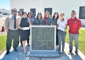 CSP-Corcoran awarded three scholarships to high school graduates. They are posing beside the plaque for the prison along with staff members and guests.