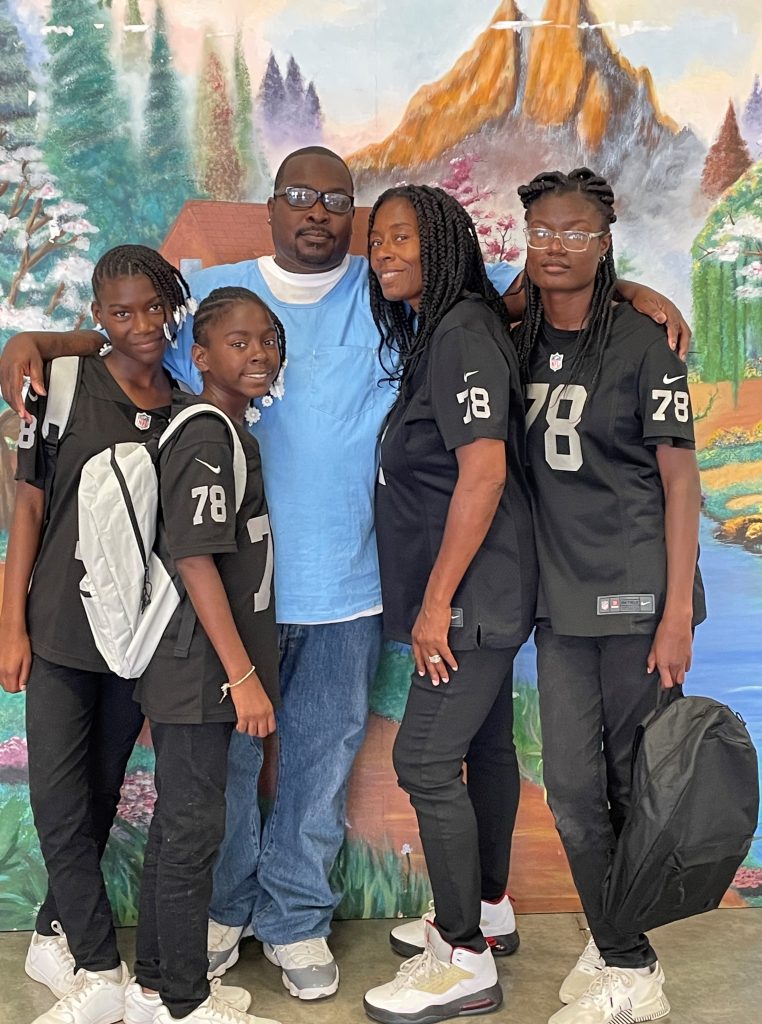 An incarcerated man with his wife and three children. The children are wearing or holding backpacks.