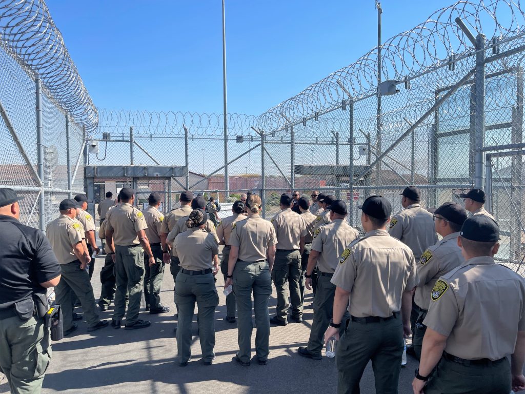 Correctional officer cadets stand in a fenced area topped by razor wire.