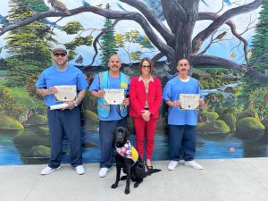Three incarcerated artists who painted a palliative care mural are shown with the CHCF warden and one of the dogs in the program called Canine Companions.
