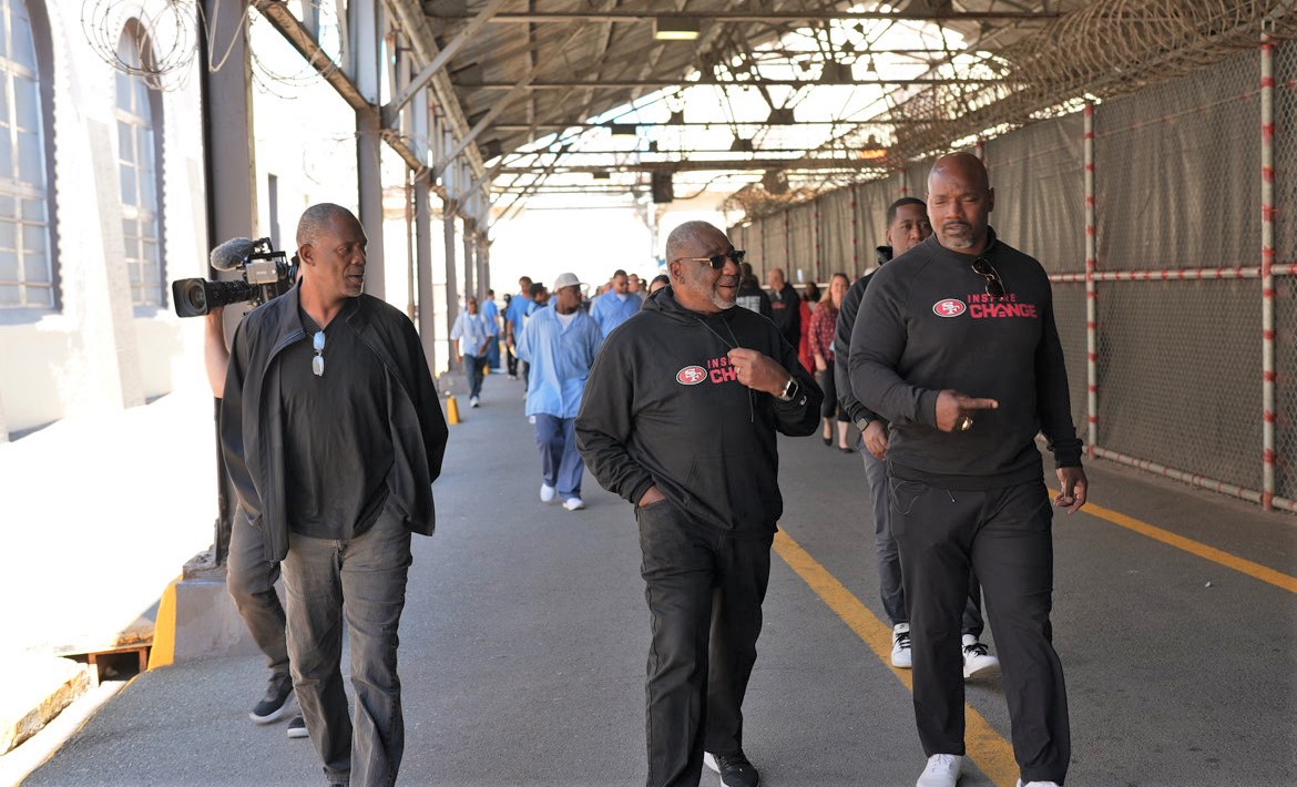 Inspire Change initiative with San Francisco 49ers walking through San Quentin Rehabilitation Center. The image shows three 49ers representatives walking along a covered path while incarcerated people can be seen behind them.