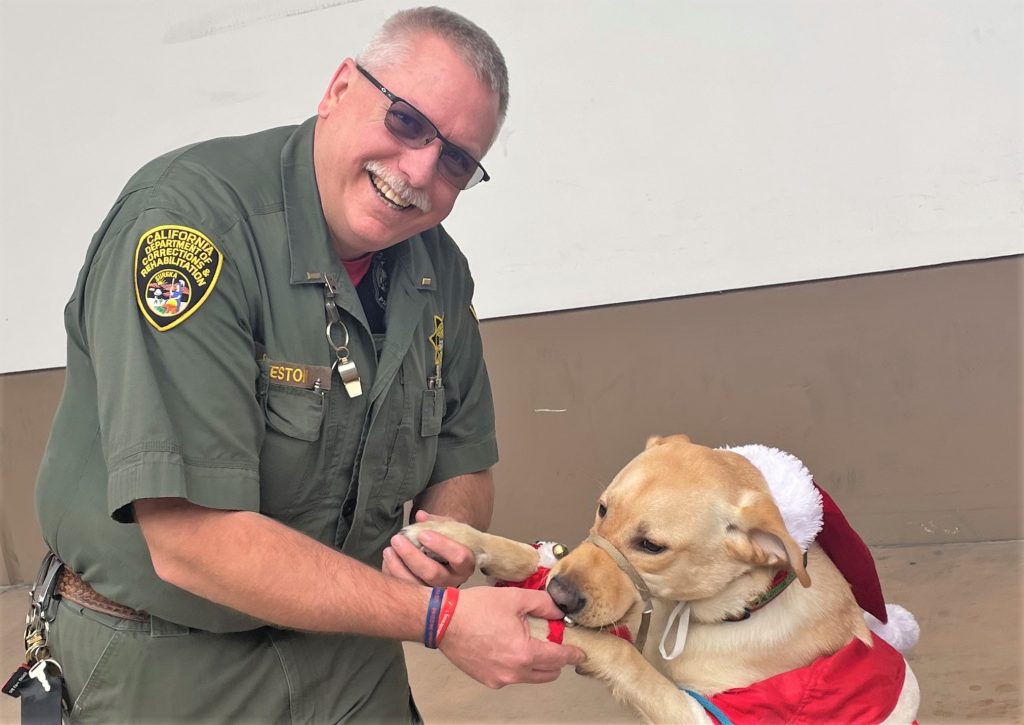 A correctional officer smiles as he holds the paws of a dog wearing a Santa outfit.