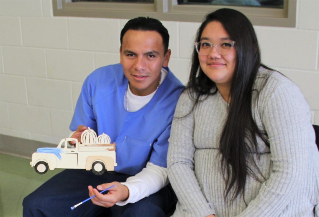 An incarcerated person holds a painting he's working on while a woman sits next to him.