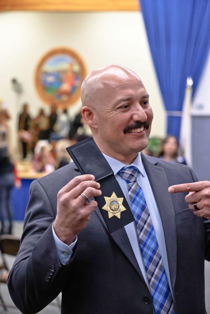 A parole agent points to his badge while smiling. 