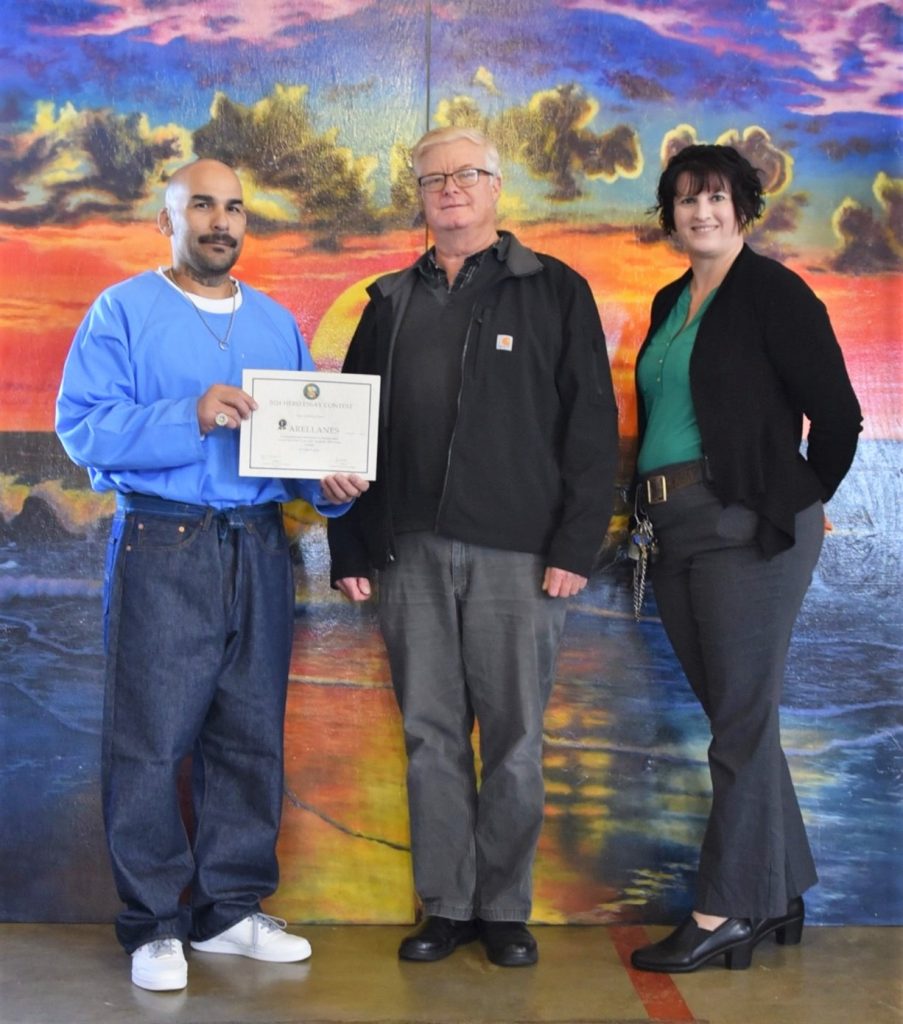 An incarcerated student holds a certificate while standing beside two staff members at Pelican Bay State Prison in Crescent City, California.