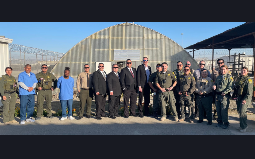Secretary Macomber with staff and incarcerated people with greenhouse in background.