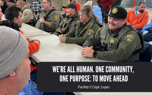 Incarcerated people and correctional staff at a CCI town hall, siting at a table, with the words "We’re all human. One community, one purpose: to move ahead." Facility C Capt. Lopez.