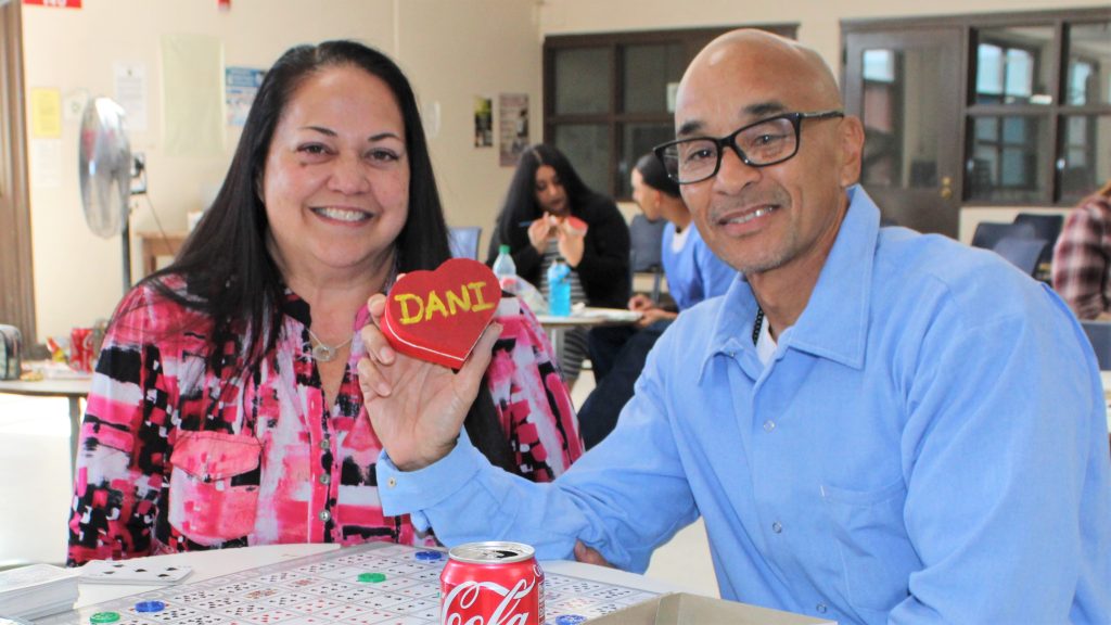 Woman holds a decorated jewelry box with the word "Dani" while sitting next to an incarcerated person at Centinela State Prison.