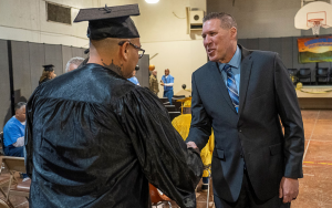 CIM warden shakes hands with an incarcerated person wearing a graduate cap and gown.