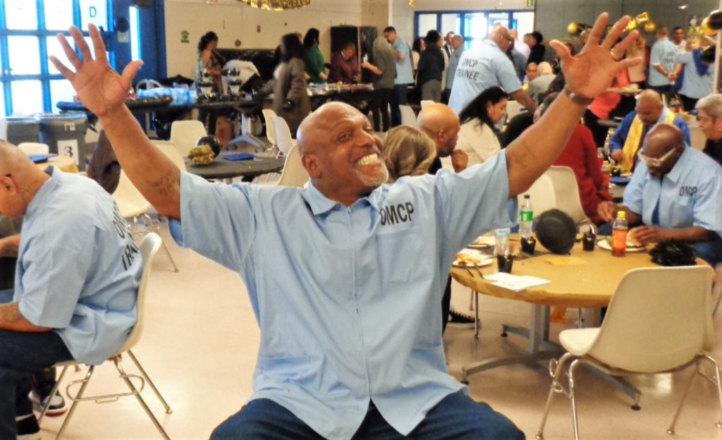 An incarcerated person smiling with arms open wide. He's wearing an OMCP shirt at the graduation at Corcoran prison.